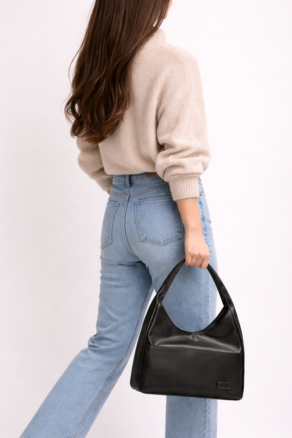 Person wearing a beige sweater and blue jeans, holding a black handbag against a light background