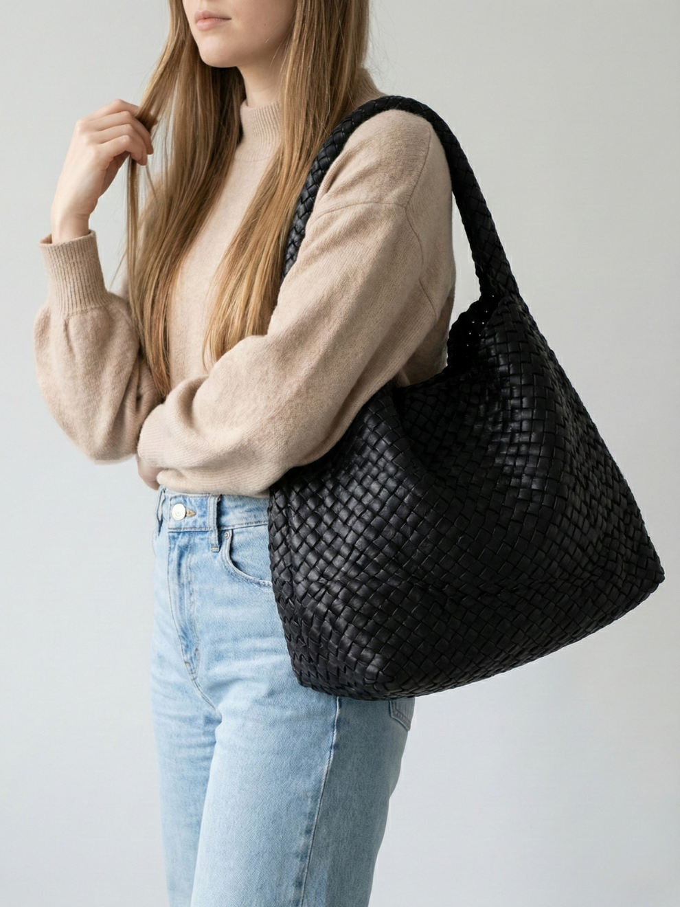Person wearing a beige sweater and light blue jeans with a black woven handbag on a plain background