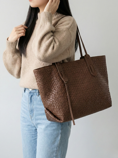 Person wearing a beige sweater and blue jeans holding a brown woven handbag against a plain background