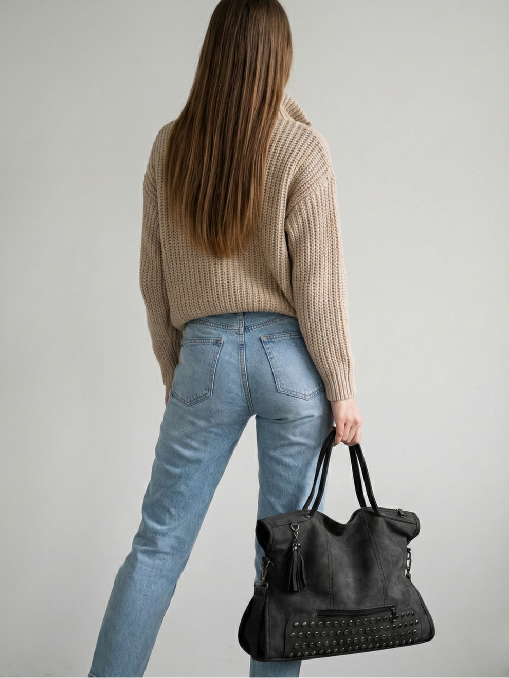 Person wearing a beige sweater and blue jeans, holding a black handbag against a plain background