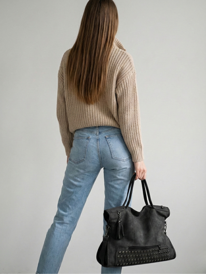 Person wearing a beige sweater and blue jeans, holding a black handbag against a plain background